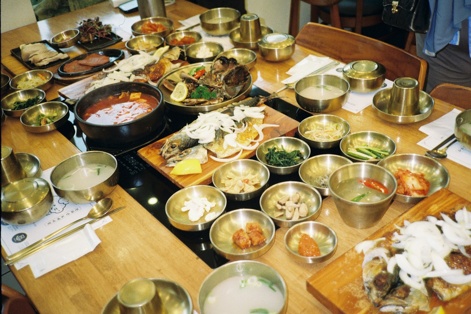 A long Korean table crowded with shared side dishes in brass bowls, a hot pot in the middle